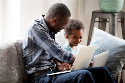a father and son sit on a couch looking at a laptop