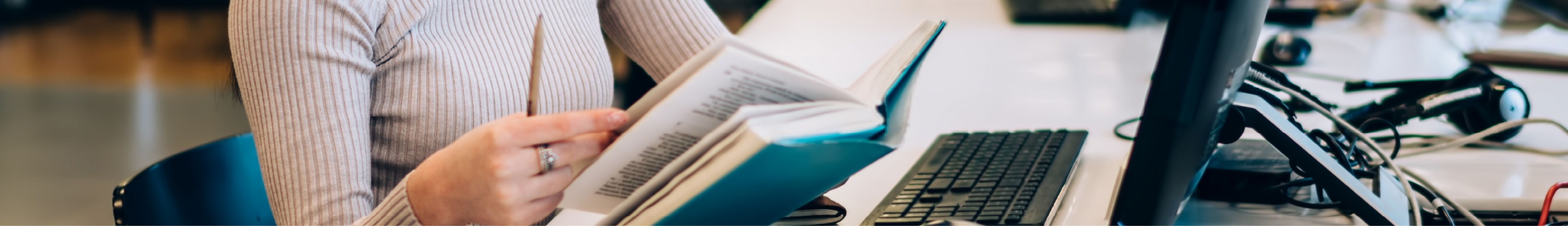 An image of a person holding a notebook open. They are sitting at a desk in front of a computer.