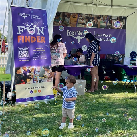 A young kid stands in front of a Fort Worth Public Library booth at Mayfest. He's holding a yellow wand toy and is surrounded by bubbles.