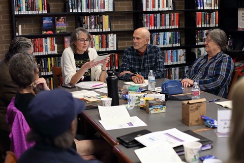 A group of people sit at a table in a library and discuss their writing.