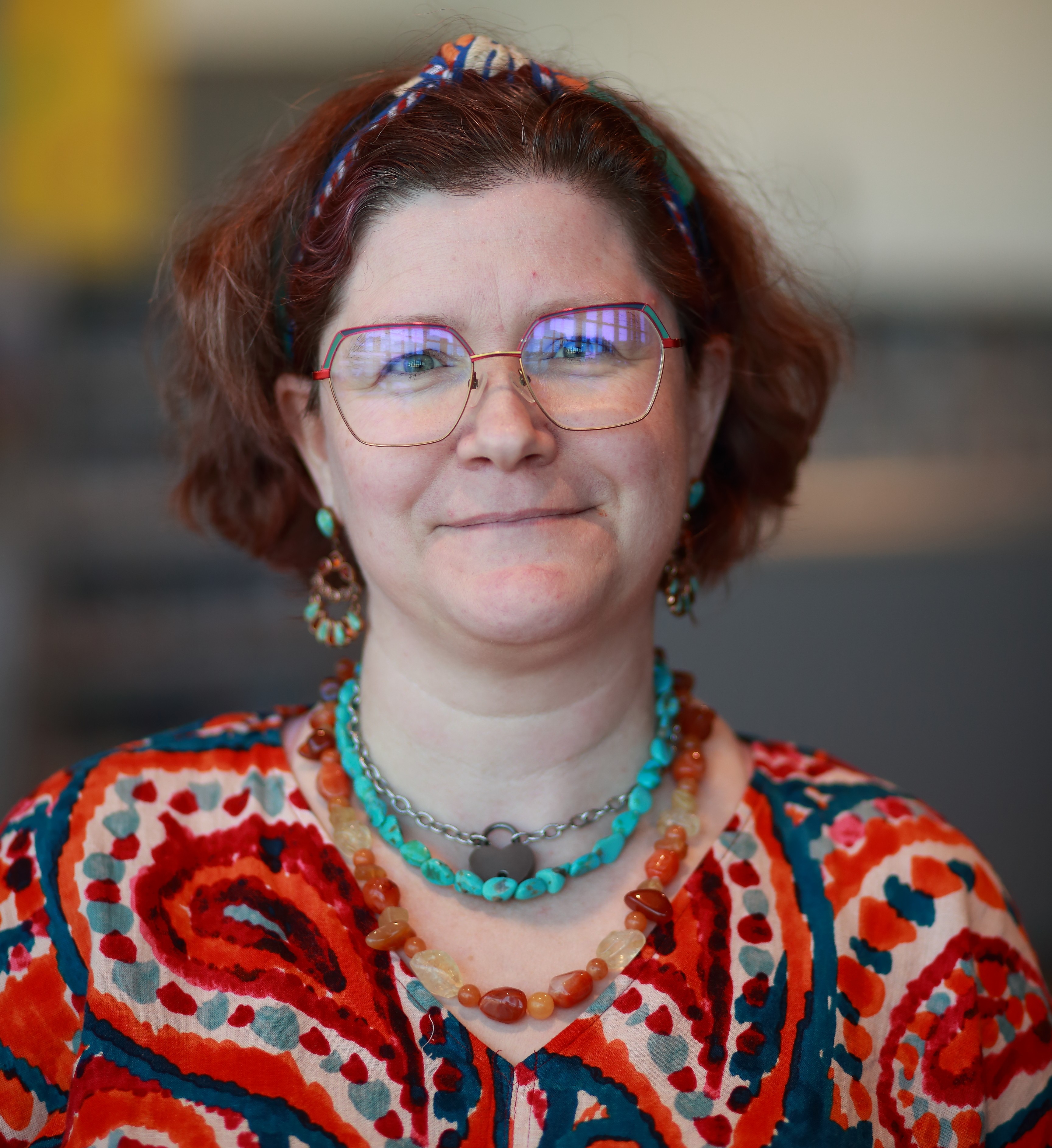 A woman poses for a photo. She is wearing red glasses and a brightly patterned shirt.