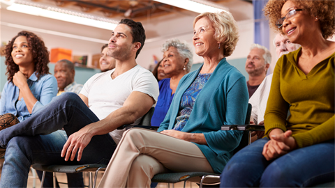 Variety of people sitting in chairs, in a meeting setting.