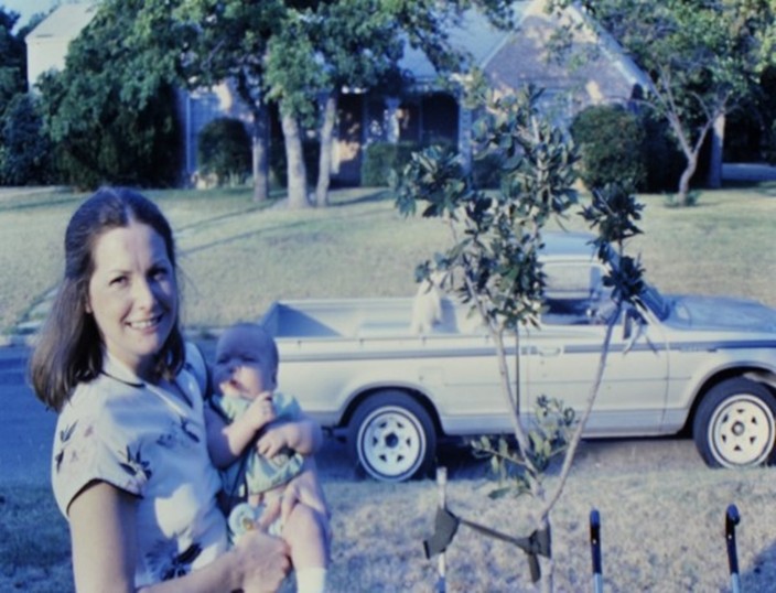 Woman Holding baby in front of newly planted tree.