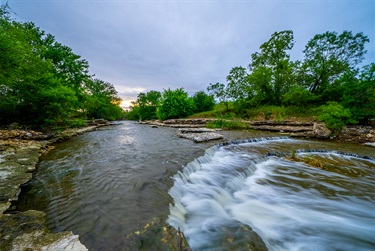 Photo of a small waterfall on Little Fossil Creek with trees along the shore and sun setting in the background