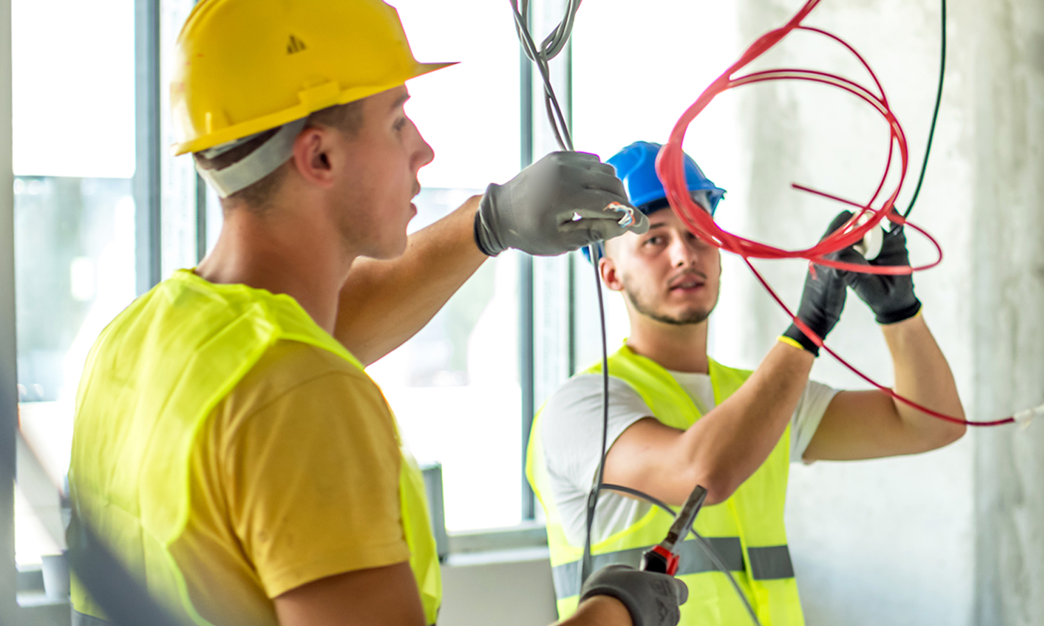 Two male electricians in hard hats and reflective vests installing ceiling wiring