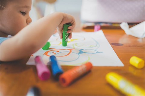 A young child sits at a wooden table coloring a white sheet of paper. Markers in various colors site around the table.