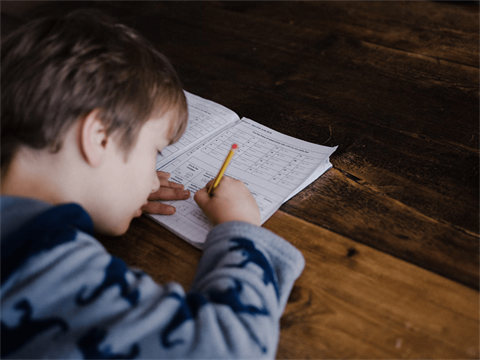 A young child sits at a wooden table and works on math problems in a notebook.