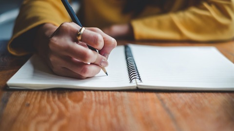 Picture of an adult wearing a yellow long-sleeve using a pen to write in a notebook.