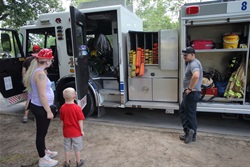 A woman and child stand on the left side of the image in front of a fire truck. The woman is wearing a fire helmet, and the child is wearing a red shirt. A fire fighter is standing on the right side of the image, learning against the fire truck.